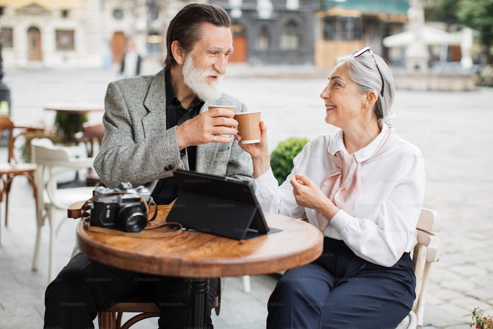 Stylish aged couple relaxing together on cafe terrace and enjoying hot coffee. Lovely wife and husband using digital tablet and retro camera outdoors.