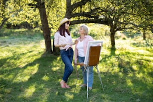 Joyful young woman in casual wear and straw hat, having fun outdoors in the garden, while painting a picture together with her mature mother. Joint activity, creativity concept.