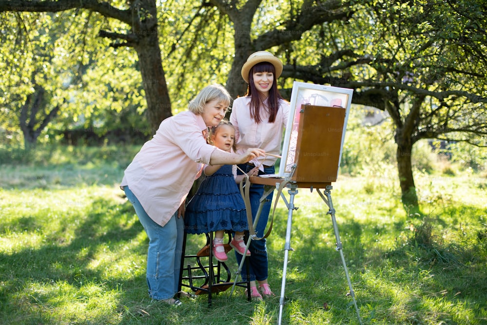 Tempo libero estivo in famiglia con hobby della pittura. Ritratto all'aperto di una famiglia di tre generazioni di donne, che trascorrono del tempo all'aperto nel parco, disegnando insieme su tela con colori.