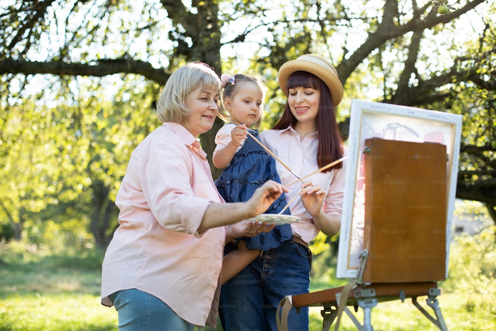 Cavalete, tela, pincéis e tintas. Lazer em família. Close up de três gerações de família, avó, mãe e menina artista, pintando juntos um quadro sobre tela ao ar livre no parque.