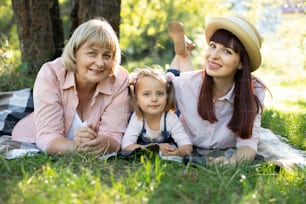 Grandmother, mother and grand daughter enjoying sunny garden holiday together outdoors, lying on green grass on blanket and smiling to camera. Leisure family lifestyle, happiness and moments.