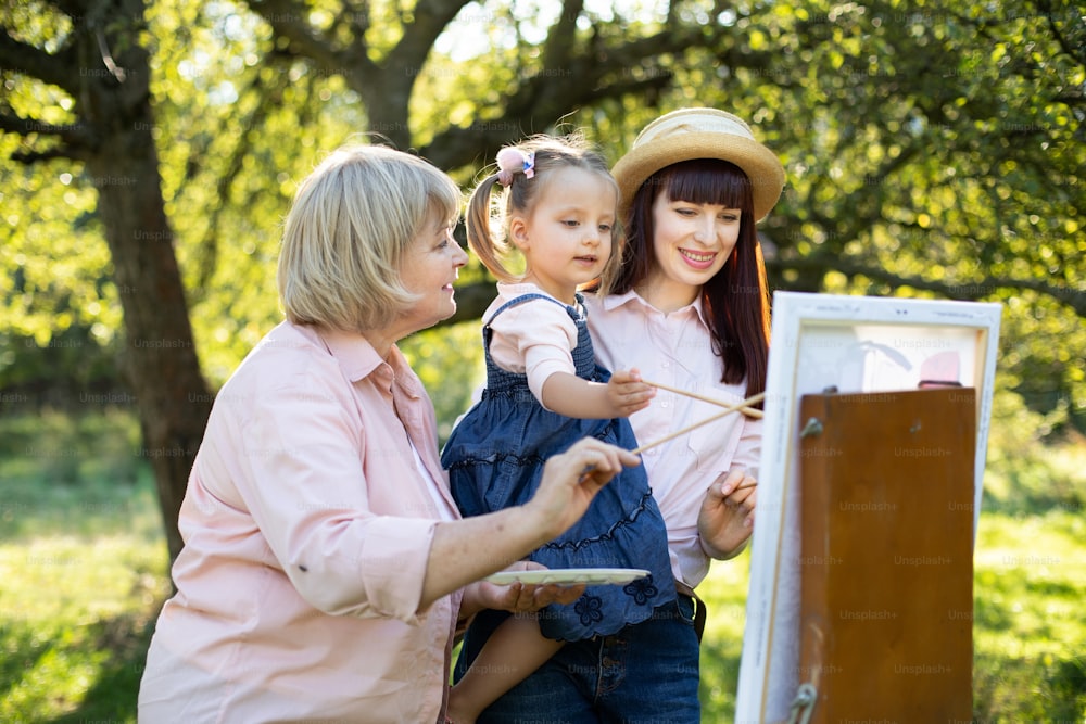 Creative and art kids development concept. Three generation women, retired grandmother, young pretty mother and cute little girl child drawing on easel in spring park