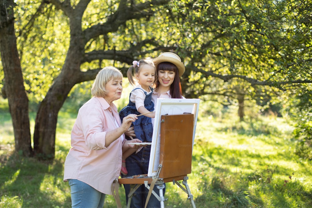 Happy family leisure, painting together. Pretty senior grandmother, young mom and little kid daughter drawing together at sunny day in beautiful summer garden on easel.