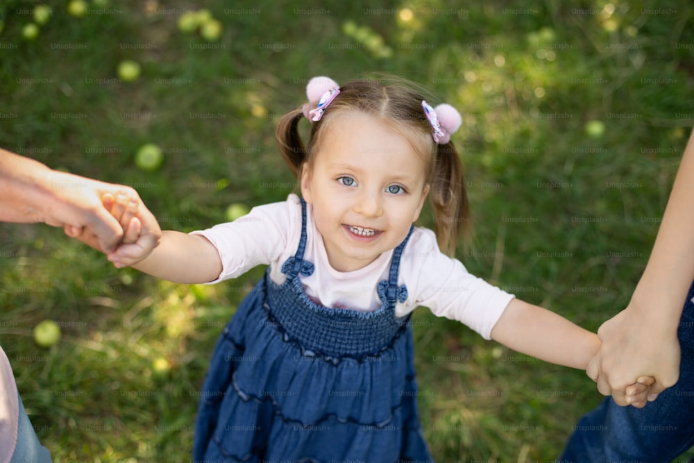 Vista superior de cerca de la adorable niña de 3 años riendo en vestido de jeans, divirtiéndose y disfrutando de su paseo al aire libre en el parque, tomada de la mano de su madre y su abuela. Paseo familiar en el jardín.