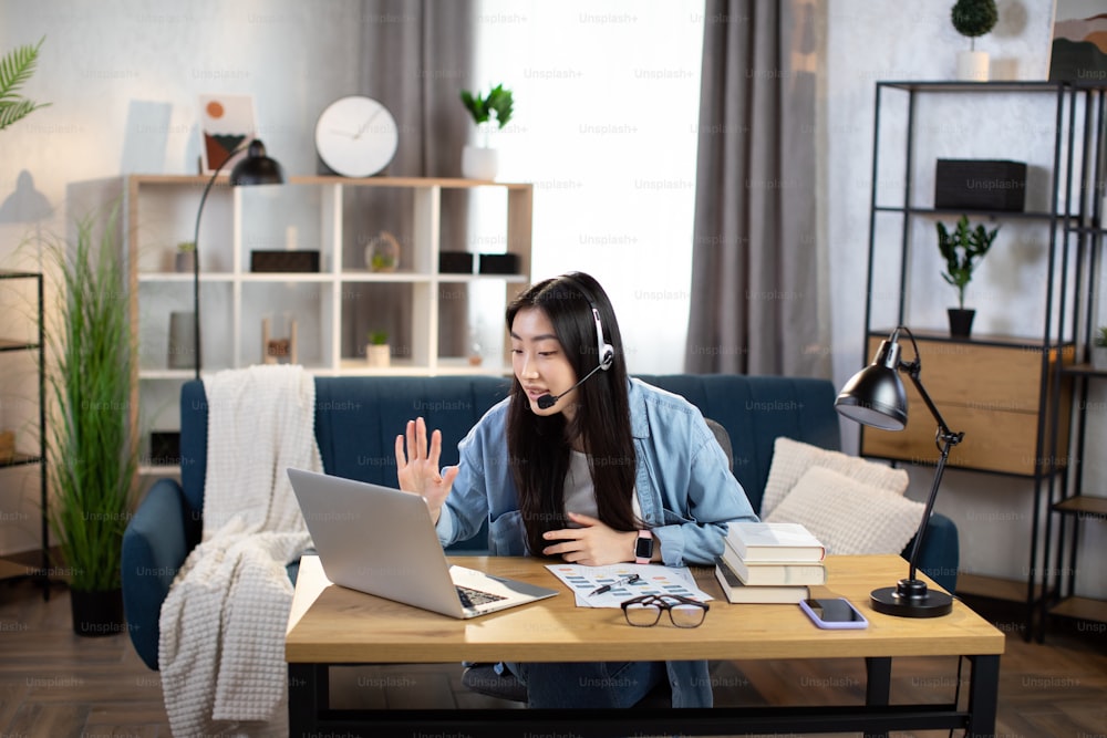 Young asian woman in headset talking and waving during video chat on modern laptop. Pretty dark-haired female having working meeting online. Work from home during pandemy