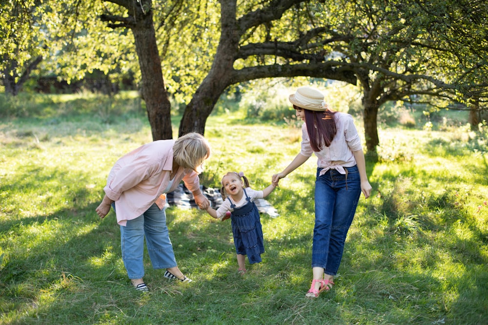 A happy Caucasian family of three generations, grandmother, daughter, and little kid girl, walking outdoors in sunny park and having fun.