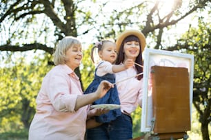 Easel, canvas, brushes and paints. Family leisure. Close up bottom view of three generation family, grandmother, mother and little girl artist, painting a picture on canvas outdoors together.