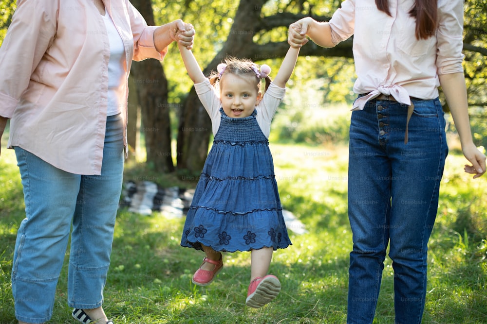 Close up shot of cute pretty little child girl, having fun outdoors and flying in the air while holding hands of her mom and grandma. Happy child walking with thei mommy and granny in the park.