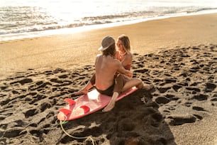 Beautiful young couple sitting on the surf board during sunset and looking at eachother with love and smile. Real people emotions. Island vibes. Summer lifestyle. Happy people having fun together, hugging, spending free time on the sandy beach.