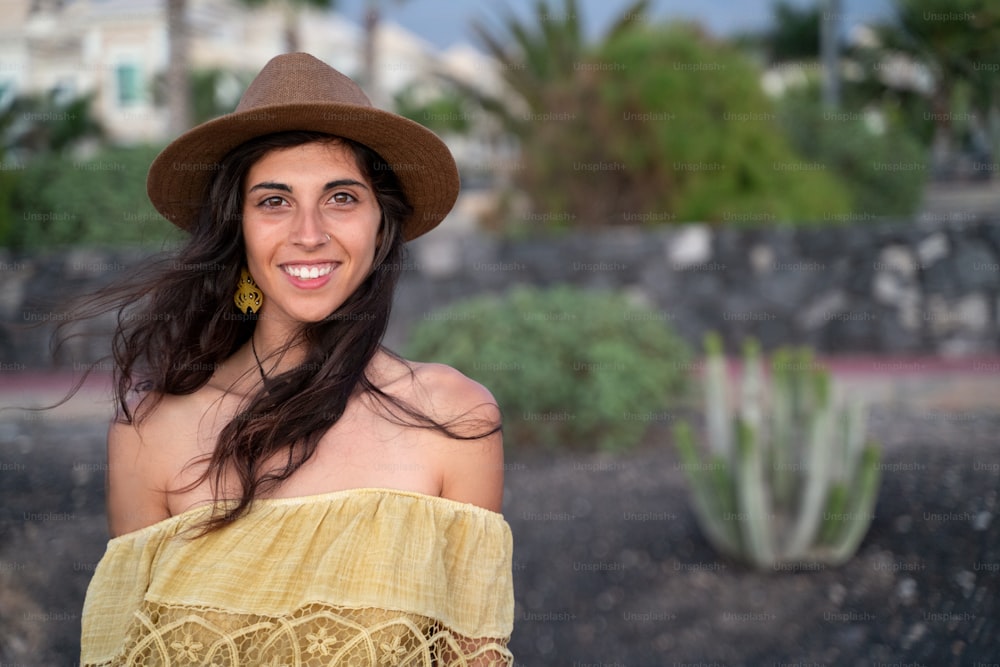 Glückliche lateinamerikanische Frau mit schönem Lächeln, die einen modischen Hut trägt und in die Kamera schaut. Junge brünette Frau, die im Sommer Urlaub genießt. Fröhliches Mädchen. Echte Menschen Emotionen.