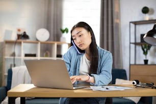 Young asian woman in casual outfit talking on mobile phone and typing on laptop while sitting at workplace. Concept of corporate communication, freelance, working online.
