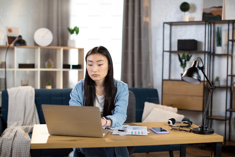 長い黒髪の美しいアジア人女性が机に座ってラップトップに入力しています。デニムシャツを着た女性が、明るいオフィスでポータブルコンピュータを使って仕事をしています。