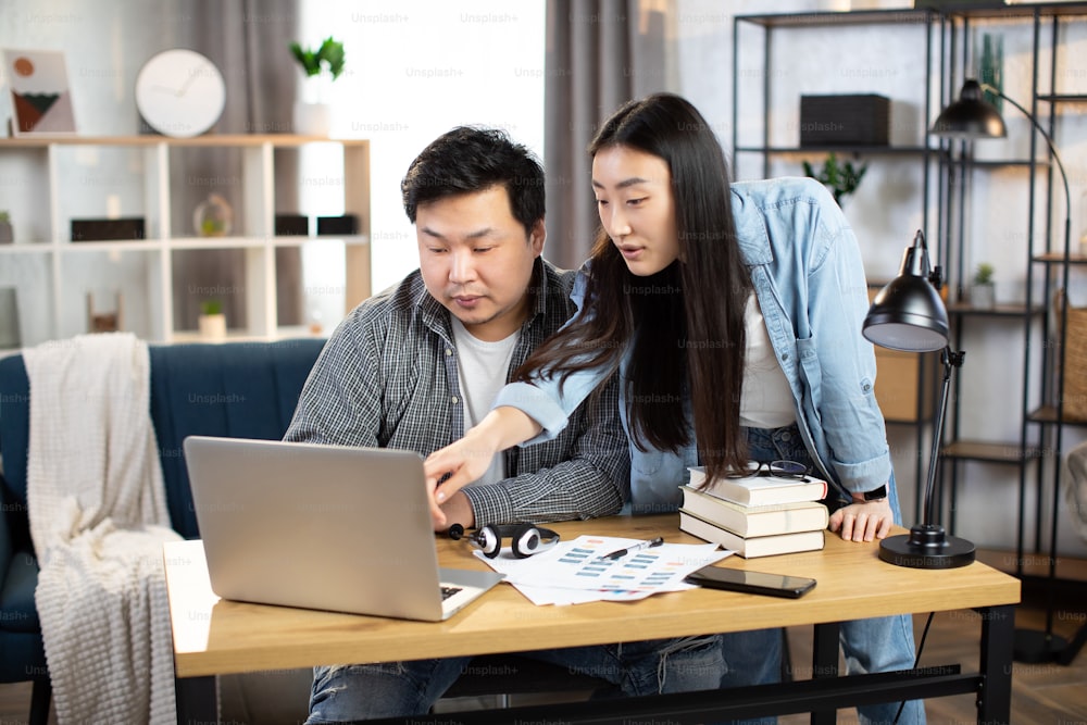 Asian man and woman in casual outfit looking at computer screen during working meeting. Young business partners cooperating at bright office or working online from home