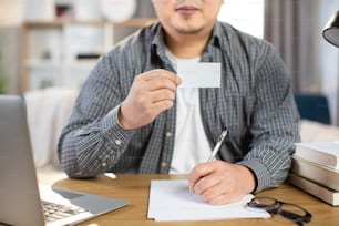 Primer plano de un joven que gana un atuendo casual sentado en el escritorio con una computadora portátil inalámbrica y que muestra una tarjeta blanca vacía. Espacio para texto en la tarjeta de visita.