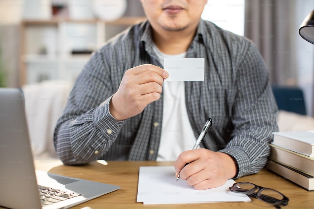 Close up of young man win casual outfit sitting at desk with wireless laptop and showing empty white card. Space for text on business card.
