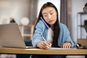 Attractive asian chinese woman talking on mobile and writing notes while sitting with modern laptop at workplace at home. Concept of gadgets and communication. Focus on hand with pen