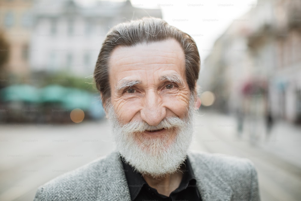 Retrato de un hombre sonriente de pelo gris vestido con ropa elegante posando en cámara en una calle de la ciudad vieja. Hombre positivo disfrutando de una jubilación feliz. Estilo de vida activo.