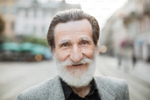 우아한 옷을 입은 웃는 회색 머리 남자의 초상화가 오래된 도시 거리에서 카메라에 포즈를 취하고 있습니다. 행복한 은퇴를 즐기는 긍정적 인 남성. 활동적인 라이프 스타일.
