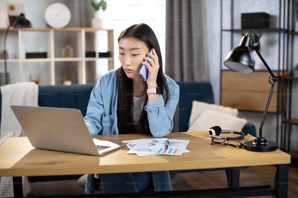 Mujer joven seria hablando en el móvil y trabajando en la computadora portátil en una oficina brillante. Morena asiática con atuendo casual usando gadgets modernos para el trabajo.