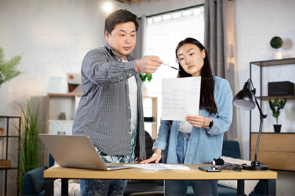 Asian man and woman designers in casual outfit, discussing project standing at bright office. Young people using modern gadgets for work. Business and technology concept.