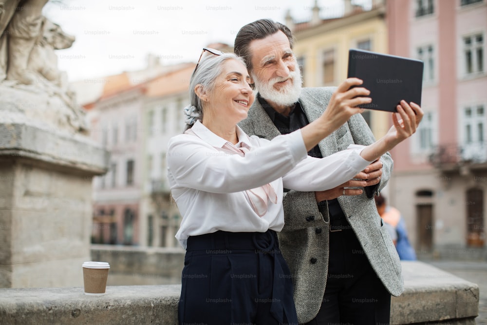 Uomo e donna anziani sorridenti che scattano selfie sulla tavoletta digitale mentre si rilassano all'aria aperta. Coppia anziana che tiene tazze di caffè. Famiglia che si gode la pensione.
