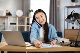Smiling asian woman writing notes while talking on mobile at bright office or home interior. Pretty female with dark hair using modern laptop and smartphone for work.