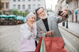 買い物袋を手に路上に立ちながら、現代のスマートフォンで自撮りをする陽気な老夫婦。家族、技術、購入のコンセプト。