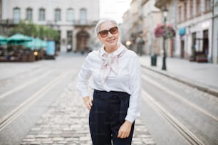 Donna adulta dai capelli grigi in abito elegante e occhiali da sole alla moda in posa e sorridente all'aperto. Strada della città sullo sfondo. Pensionato felice.