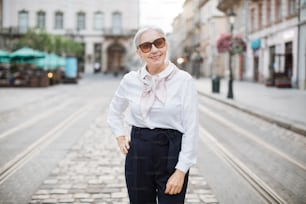 Adult grey-haired woman in elegant outfit and trendy sunglasses posing and smiling outdoors. City street on background. Happy pensioner.