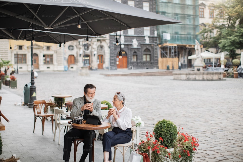 Familia madura feliz bebiendo café en la terraza de la cafetería. Hombre y mujer con estilo usando tableta y cámara retro. Relajarse al aire libre después de ir de compras.