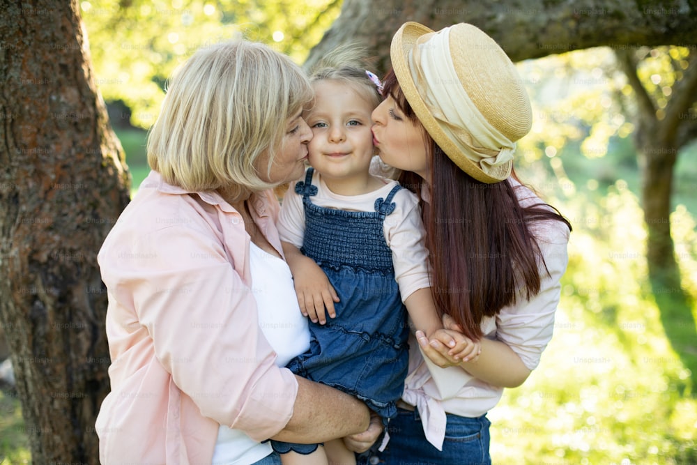 Close up picture of lovely three generation family in the park. Happy little girl smiling to camera, while her affectionate mother and senior grandmother kissing her cheeks.