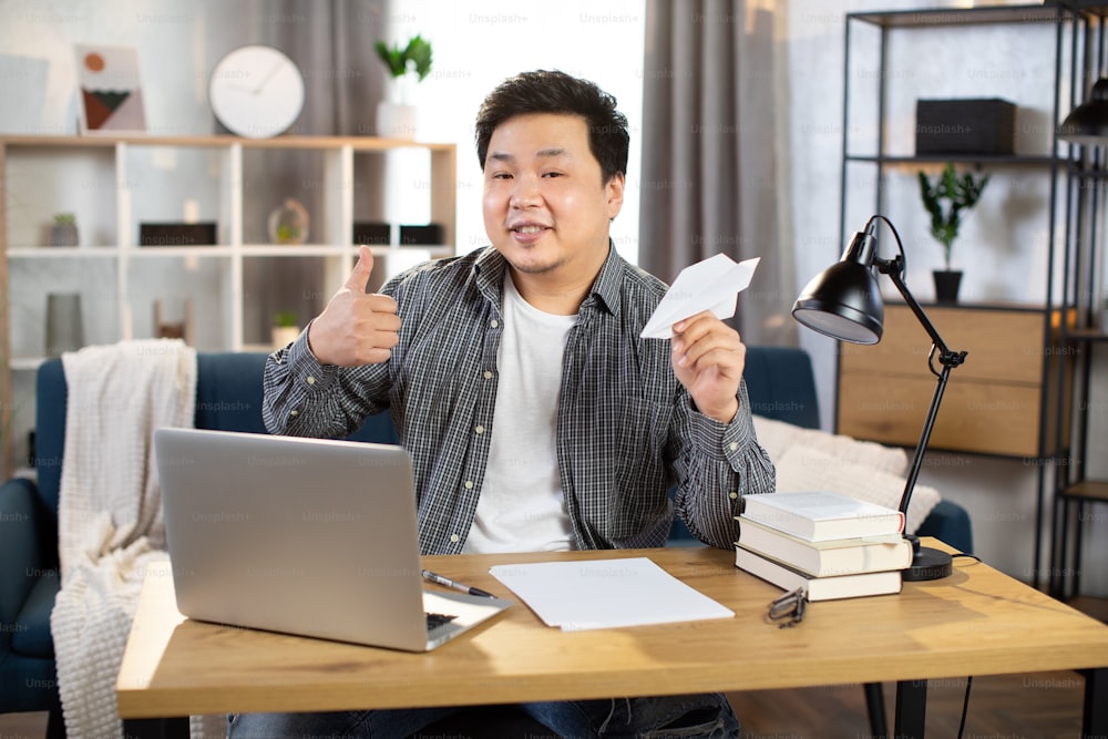 Handsome young asian chinese businessman, sitting at desk with wireless laptop, showing thumb up and playing with paper plane. Young man in casual clothes relaxing at work at cozy home living room
