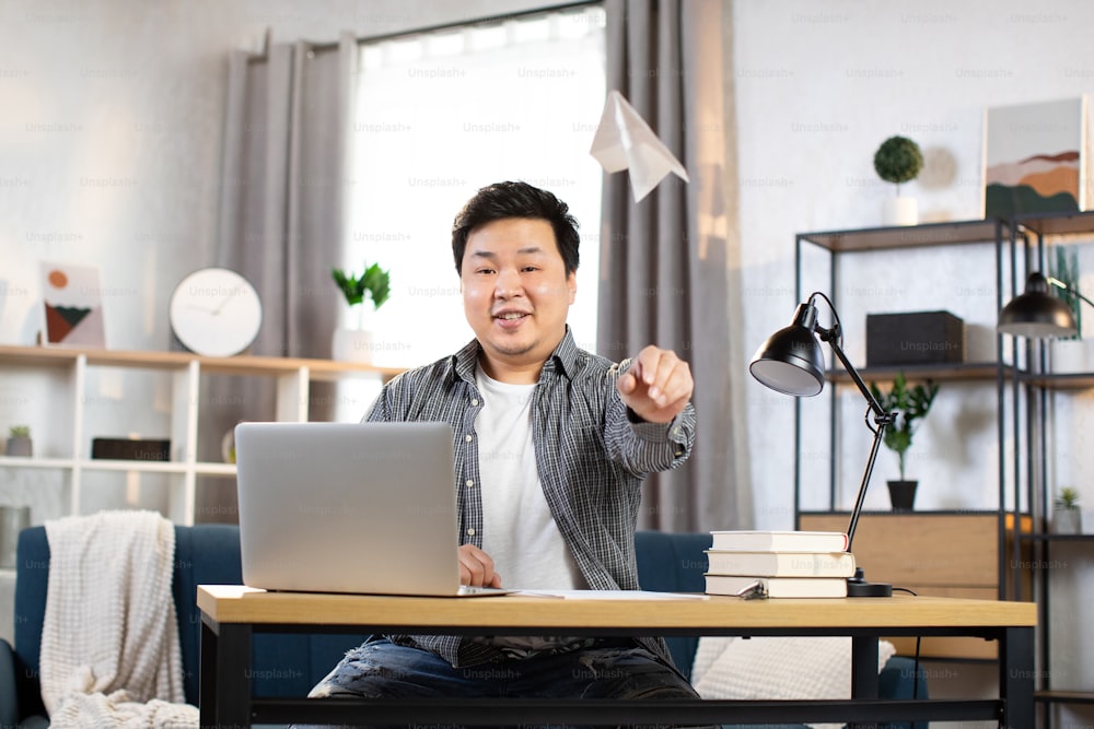 Cheerful male worker in casual outfit playing with paper plane at workplace at cozy room at home. Asian man sitting at table with wireless laptop and documents. Distance work