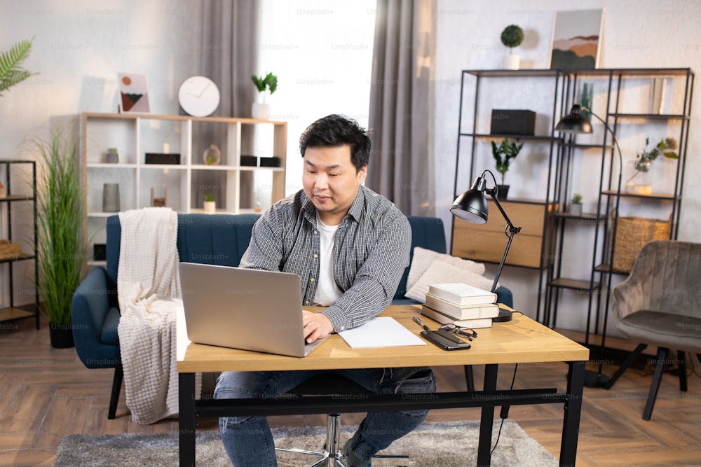 Confident asian man in casual wear typing on laptop while sitting at workplace at cozy living room at home. Busy office worker using modern gadgets for work.