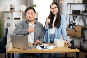 Positives asiatisches Paar, Männer und Frauen in Freizeitkleidung, posieren im hellen Büro oder zu Hause mit modernen Geräten auf dem Tisch und zeigen Daumen hoch. Konzept der Kooperation, Menschen und Technologie.
