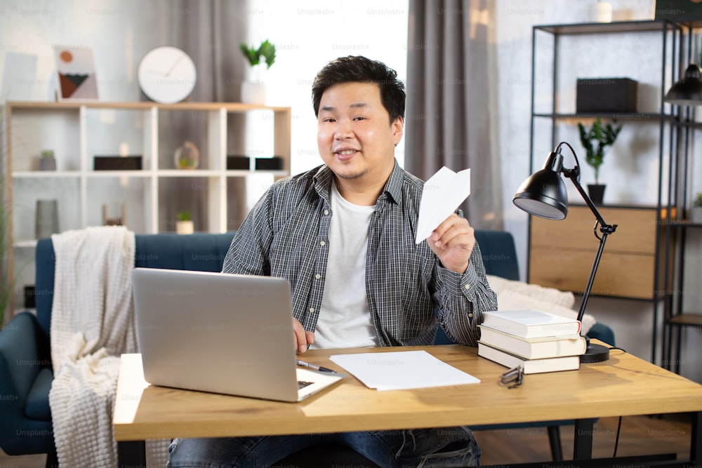 Smiling asian chinese freelancer sitting at desk with wireless laptop and playing with paper plane. Young man in casual clothes relaxing at work.