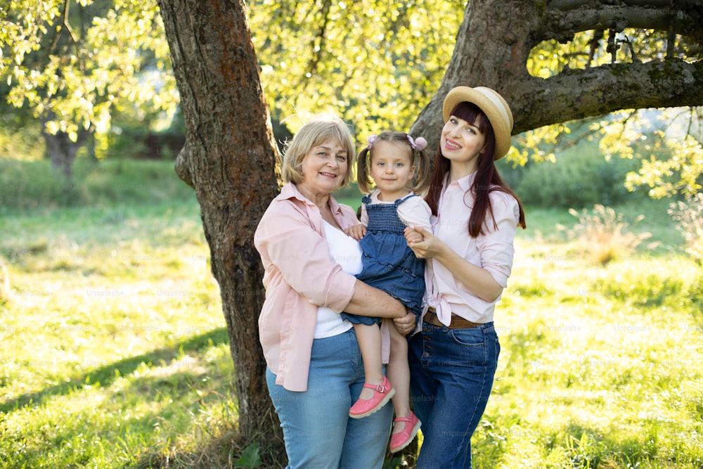 Portrait lumineux en plein air d’une famille multigénérationnelle joyeuse et heureuse, d’une grand-mère souriante, d’une jeune mère et d’une petite fille mignonne de 3 ans, posant à la caméra dans le jardin, debout sous un grand arbre.