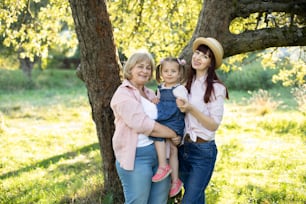 Outdoor bright portrait of happy cheerful multi generation family, smiling grandmother, young mother and little cute 3 years old girl, posing to camera in the garden, standing under big tree.