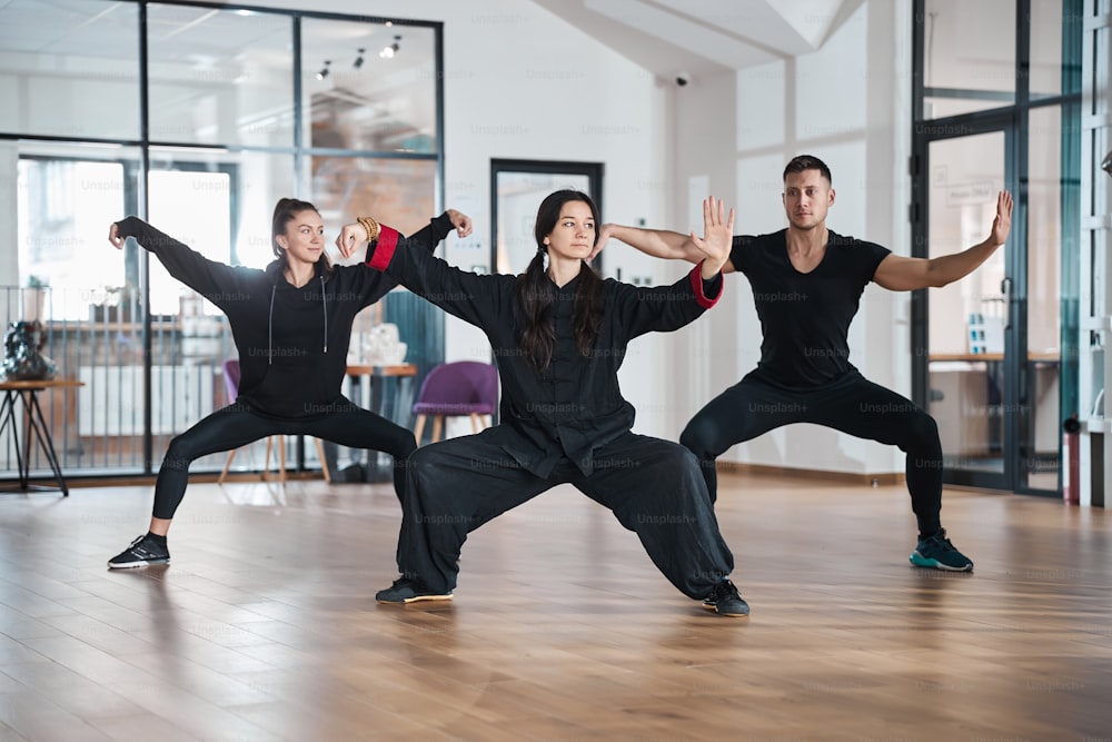 Martial arts mentor in black uniform standing with spread legs and arms before two people with opened left palm