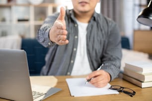 Close up of likable asian man in casual clothes looking at camera with outstretched hand for greeting. Young male freelancer sitting at table in home interior and using modern laptop.