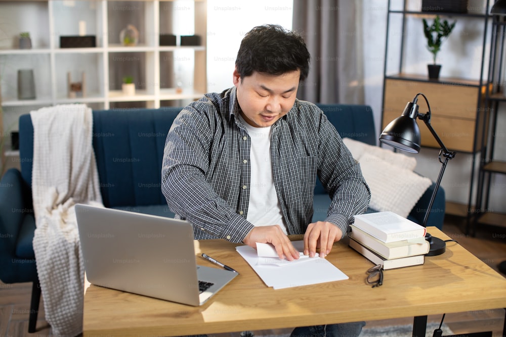 Happy asian korean man in casual wear sitting at table with laptop and making plane from paper. Relaxation and fun at work. Freelance concept.