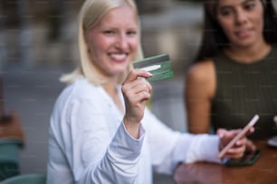 Duas mulheres no café. Mulher usando cartão de crédito e telefone celular. O foco está no cartão de crédito.