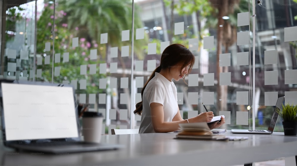 Concentre a empresária sentada no escritório moderno trabalhando com laptop e escrevendo algo o notebook.