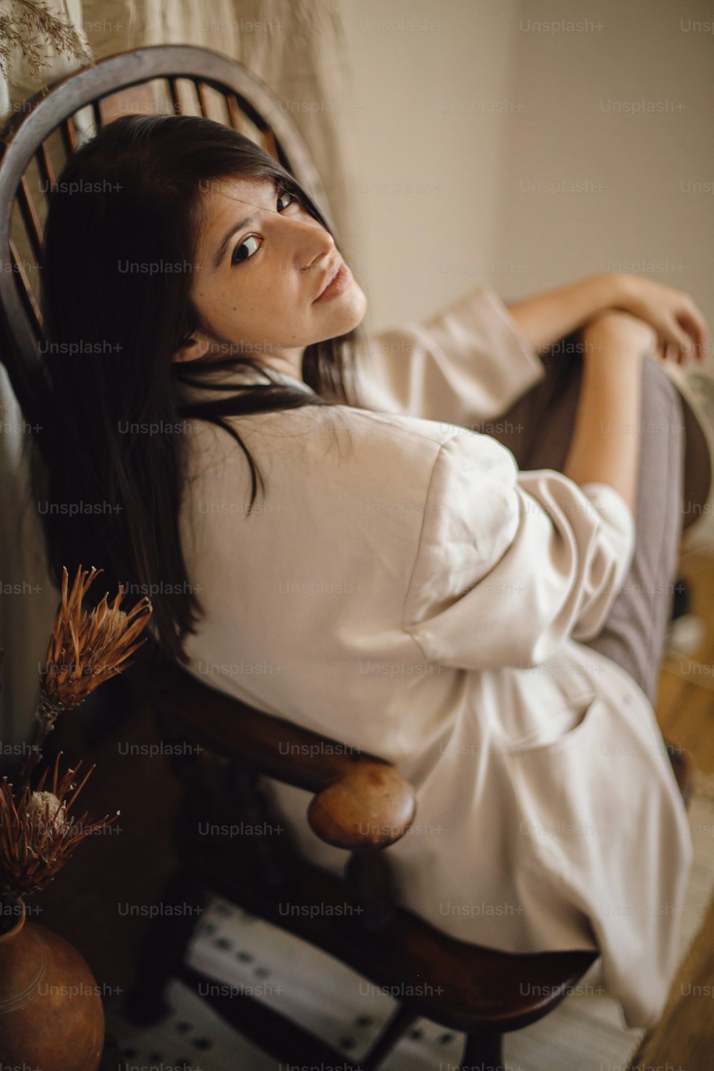 Beautiful stylish woman in suit sitting on retro wooden chair on background bohemian style room with dried grass and flowers in vase. Young fashionable female posing in pastel boho interior