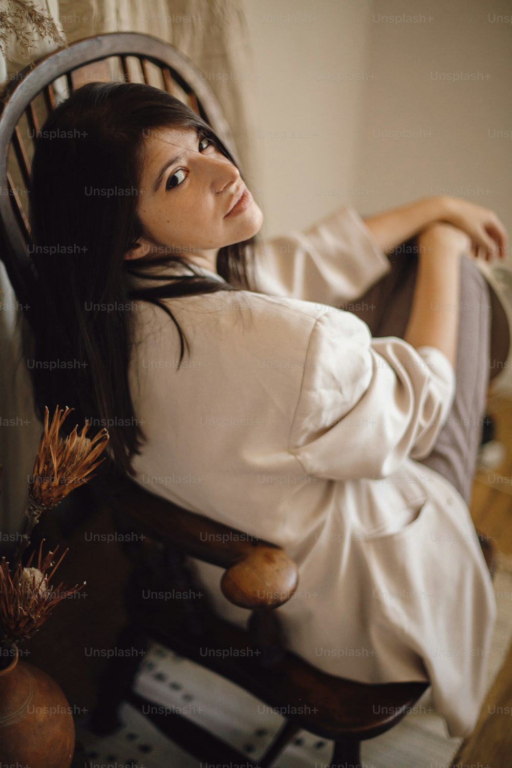 Schöne stilvolle Frau im Anzug, die auf einem Retro-Holzstuhl im böhmischen Stil mit getrocknetem Gras und Blumen in der Vase sitzt. Junge modische Frau, die im pastellfarbenen Boho-Interieur posiert