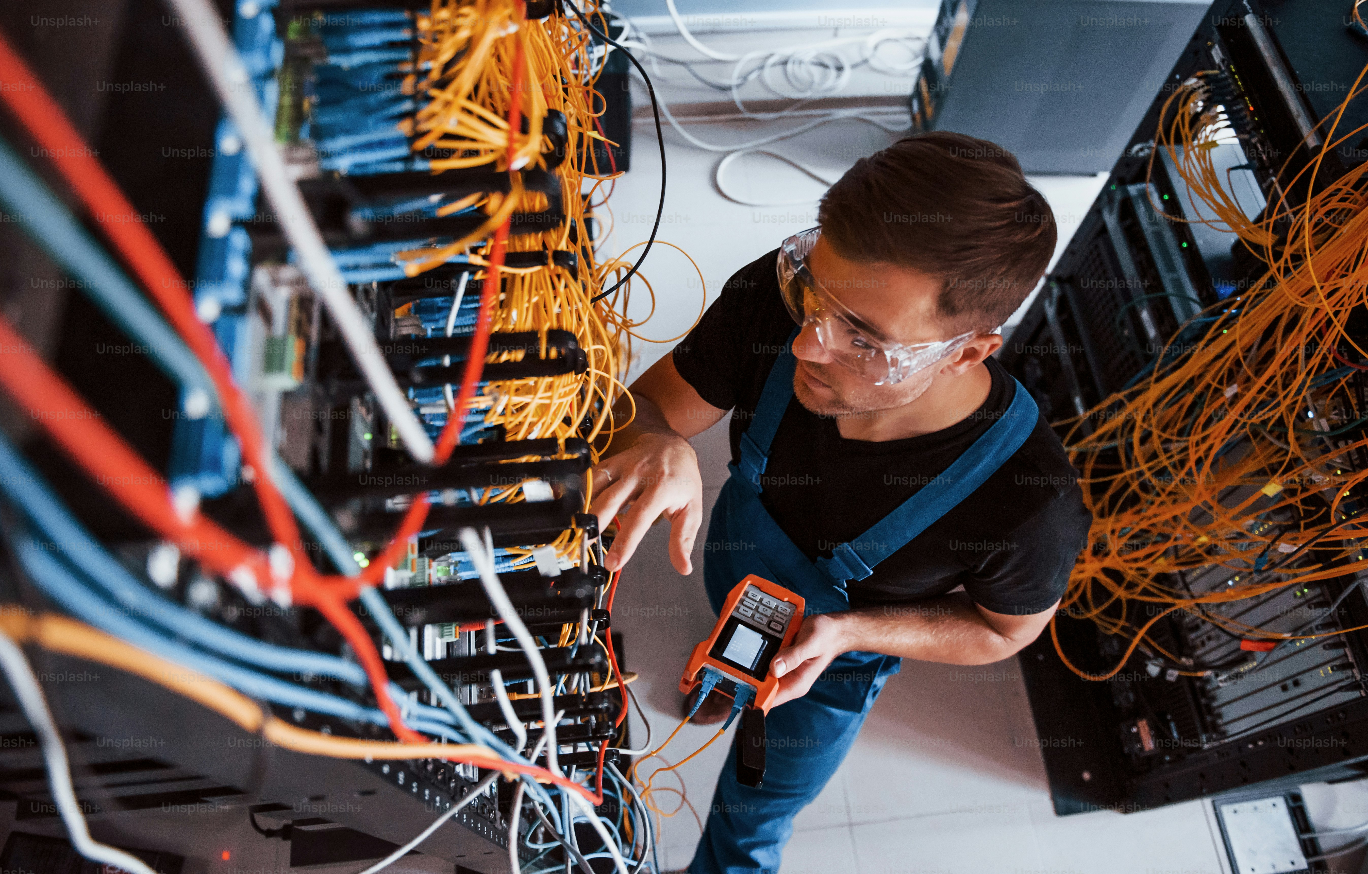 Draufsicht eines jungen Mannes in Uniform mit Messgerät, das mit Internetgeräten und Kabeln im Serverraum arbeitet.