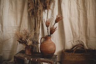 Dry protea flowers in clay vase, pampas grass, wicker basket on boho rug on background of beige linen cloth. Atmospheric bohemian style room details. Boho decor for wedding or in studio