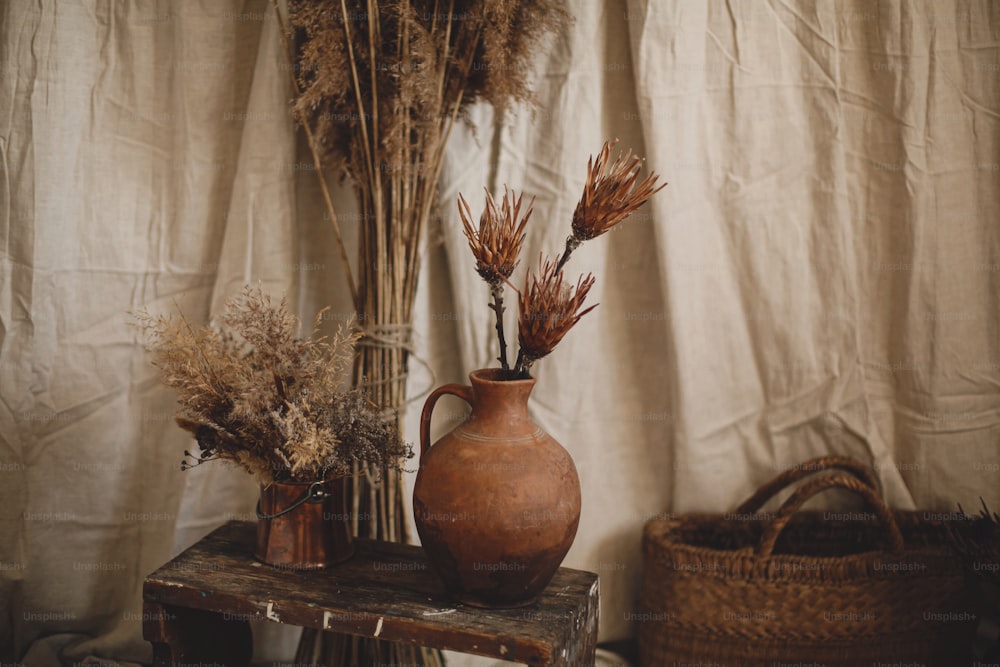 Dry protea flowers in clay vase, pampas grass, wicker basket on boho rug on background of beige linen cloth. Atmospheric bohemian style room details. Boho decor for wedding or in studio