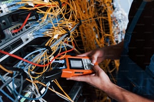 Young man in uniform with measuring device works with internet equipment and wires in server room.