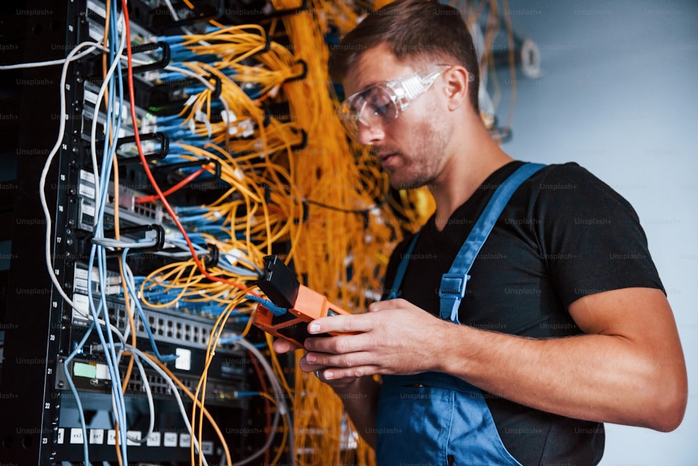 Young man in uniform with measuring device works with internet equipment and wires in server room.